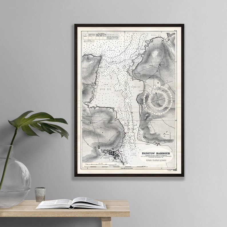 Framed map of Padstow Harbour on a wall above a wooden table with an open book and plant.