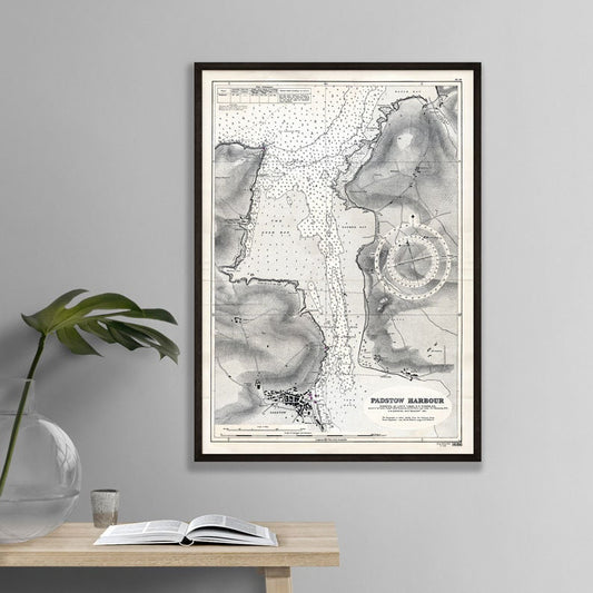 Framed map of Padstow Harbour on a wall above a wooden table with an open book and plant.
